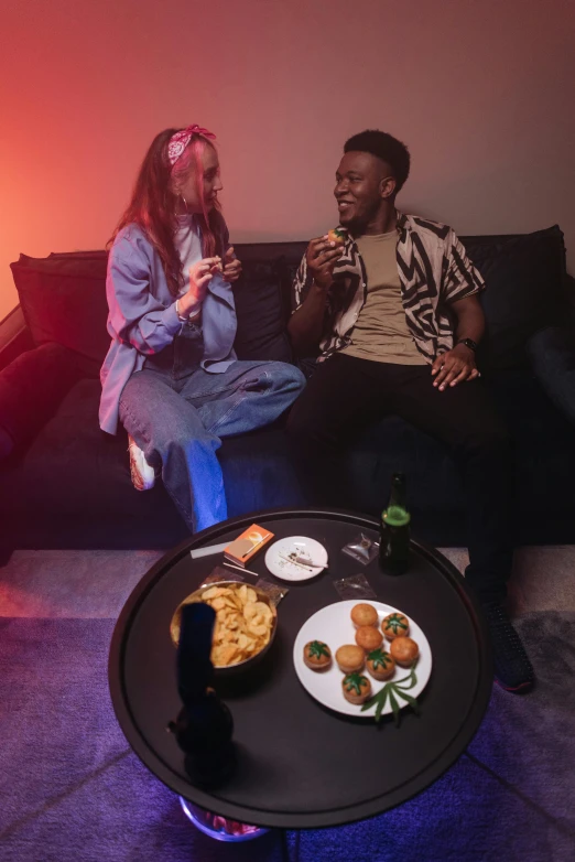 two people sitting down on a couch with food on the table