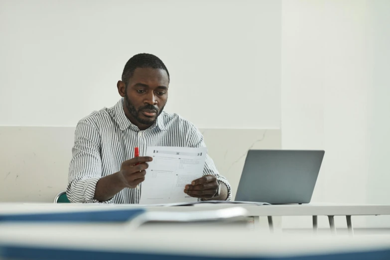 a man holding up some paper while looking at his laptop