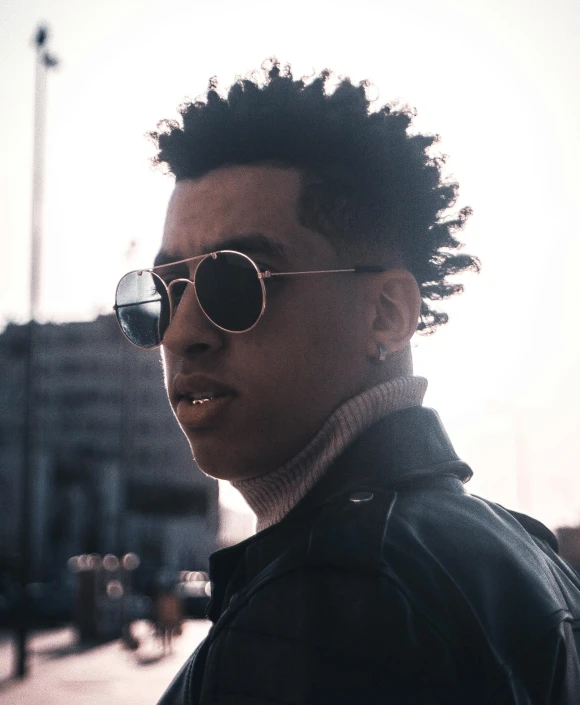 a black male with a black leather jacket, sunglasses, and a black jacket is standing on the street corner