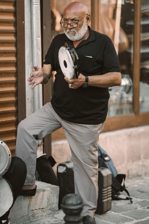 an older man in grey pants and a black t - shirt, leaning on a pillar and holding a tambour, playing a song