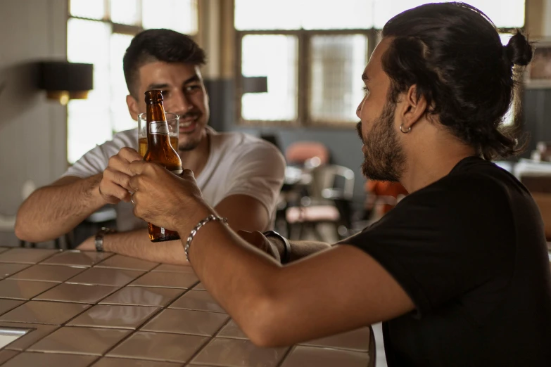 two men at a bar clinking each other with beer bottles