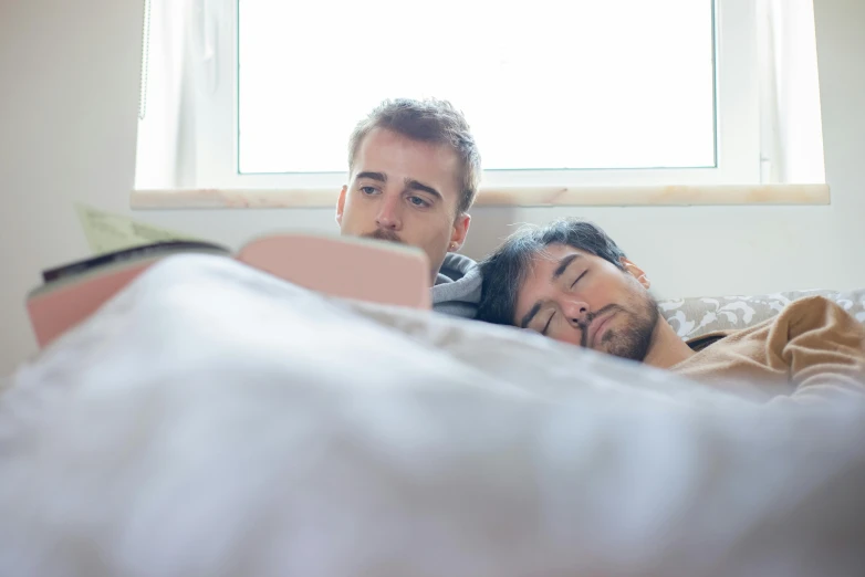 two men lay in bed looking at an open book