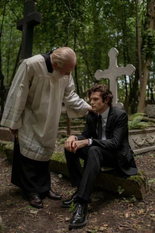 an old man standing next to a young man sitting on a bench in a cemetery