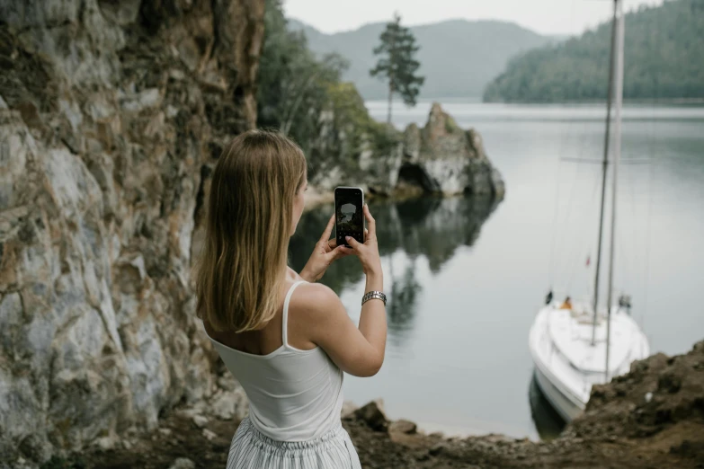 a woman standing by a body of water taking a picture of a boat