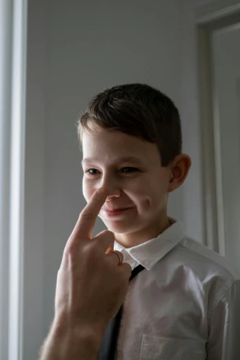 a small boy wearing a dress shirt and tie holds his finger up to his nose as he smiles
