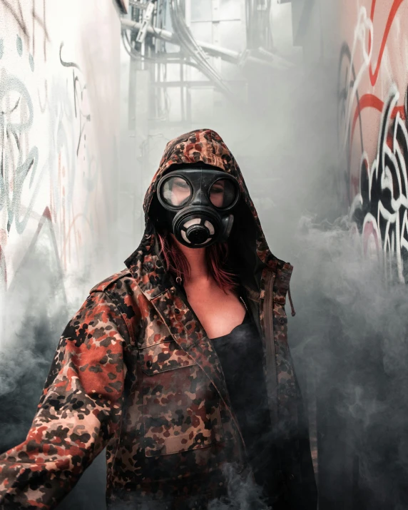 a woman with goggles, wearing an oversize, hood and jacket, stands against graffiti covered walls