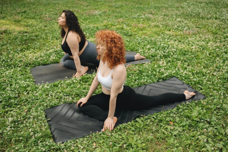 two women in yoga clothes sit in a grassy area on black exercise mats