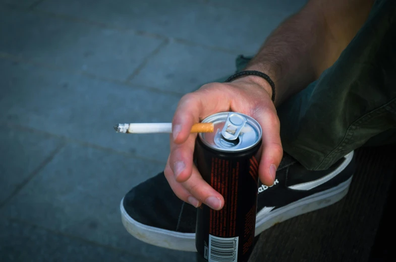 man with a cigarette and an energy drink sitting on a bench