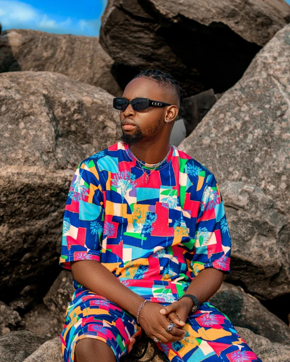 the man is sitting on the rocks wearing a multi colored shirt