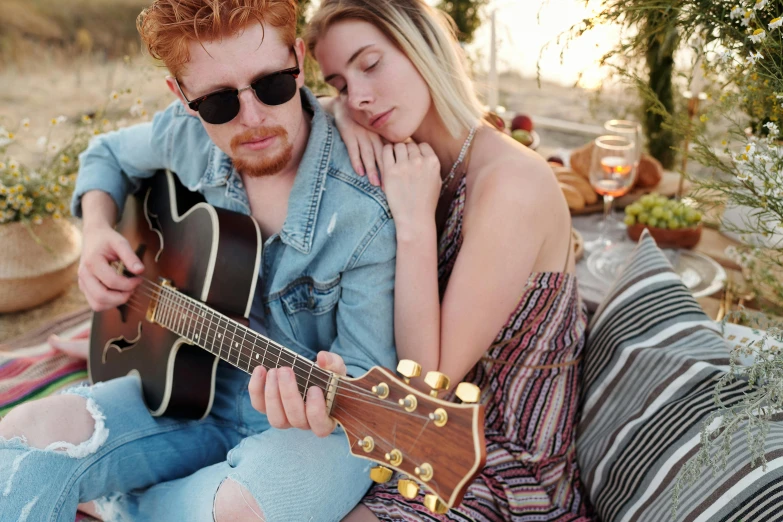 a man and woman sitting on a blanket, holding acoustic guitars