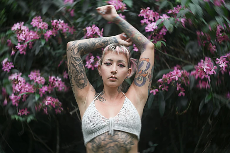 a tattooed girl wearing a white top standing with purple flowers in the background