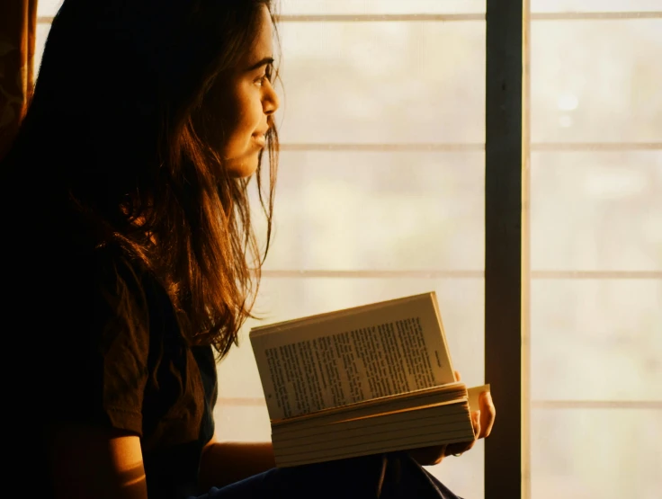 a girl reads a book next to a window