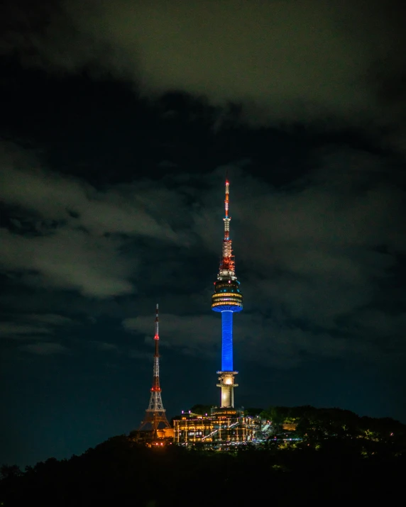 the view from the top of the hill at night, with some very colorful lights on the spire