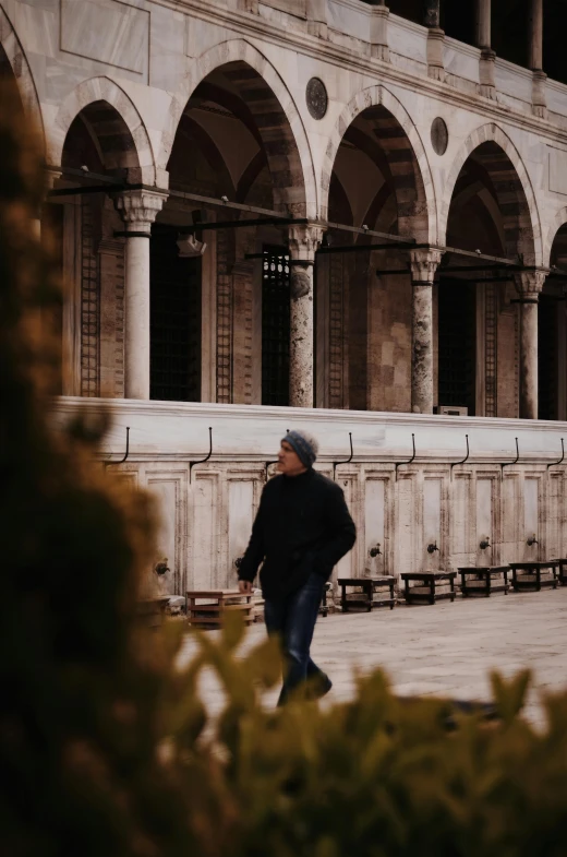 a man in black coat and hat walking next to building with arches