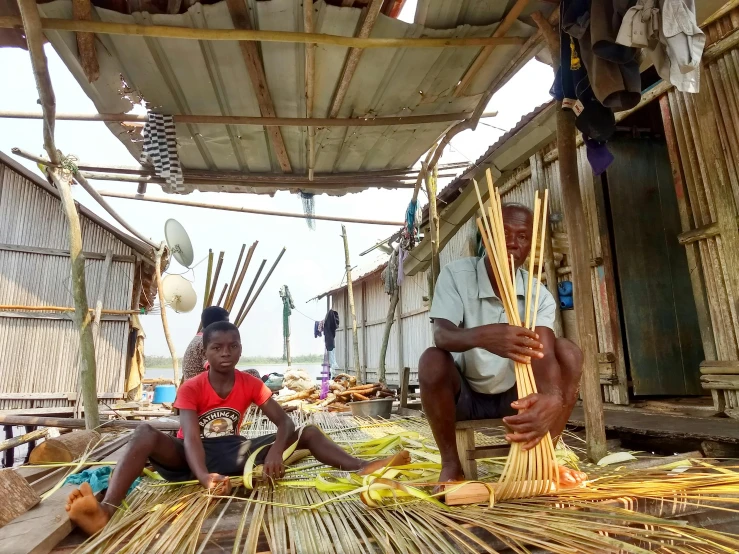 two boys sitting in front of some bamboo huts with straw weaving