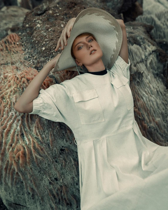 a woman in a hat and dress leaning against a large rock