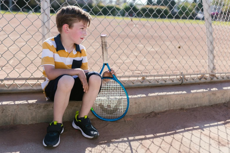 a boy sits and holds a tennis racket by the fence