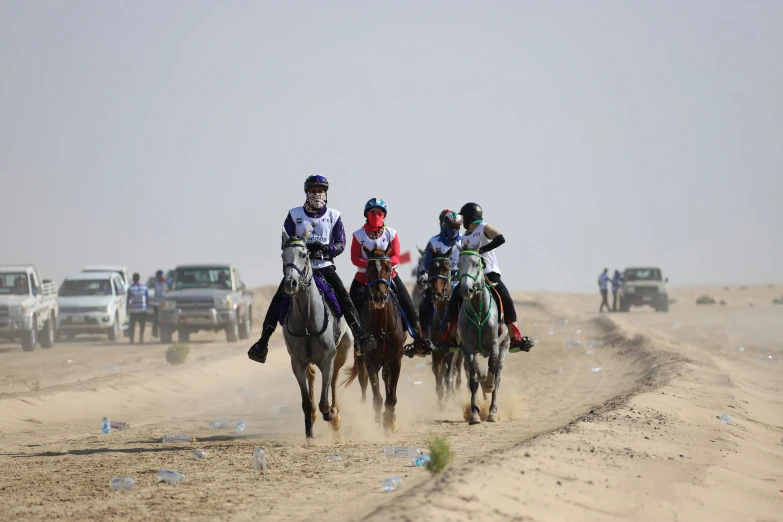 four jockeys ride horses through the desert by some trucks