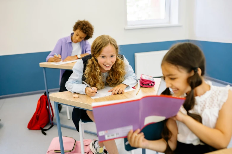 two girls sit at desks smiling and reading