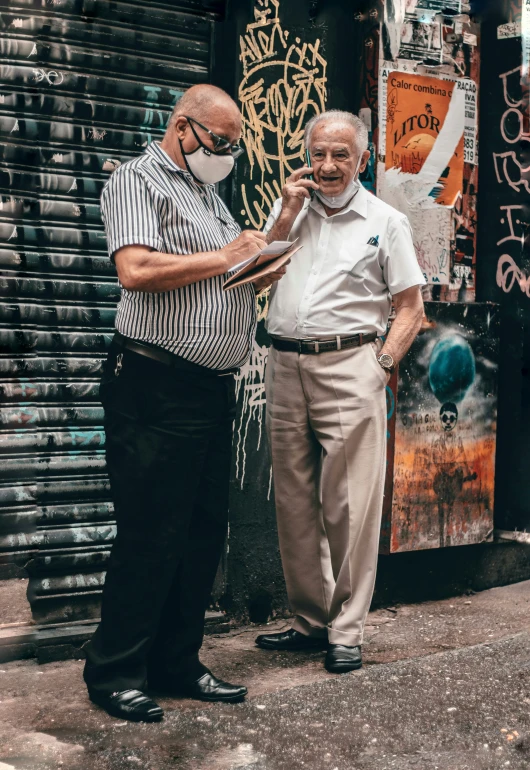 two older men with masks on stand next to each other in front of graffiti - covered doors