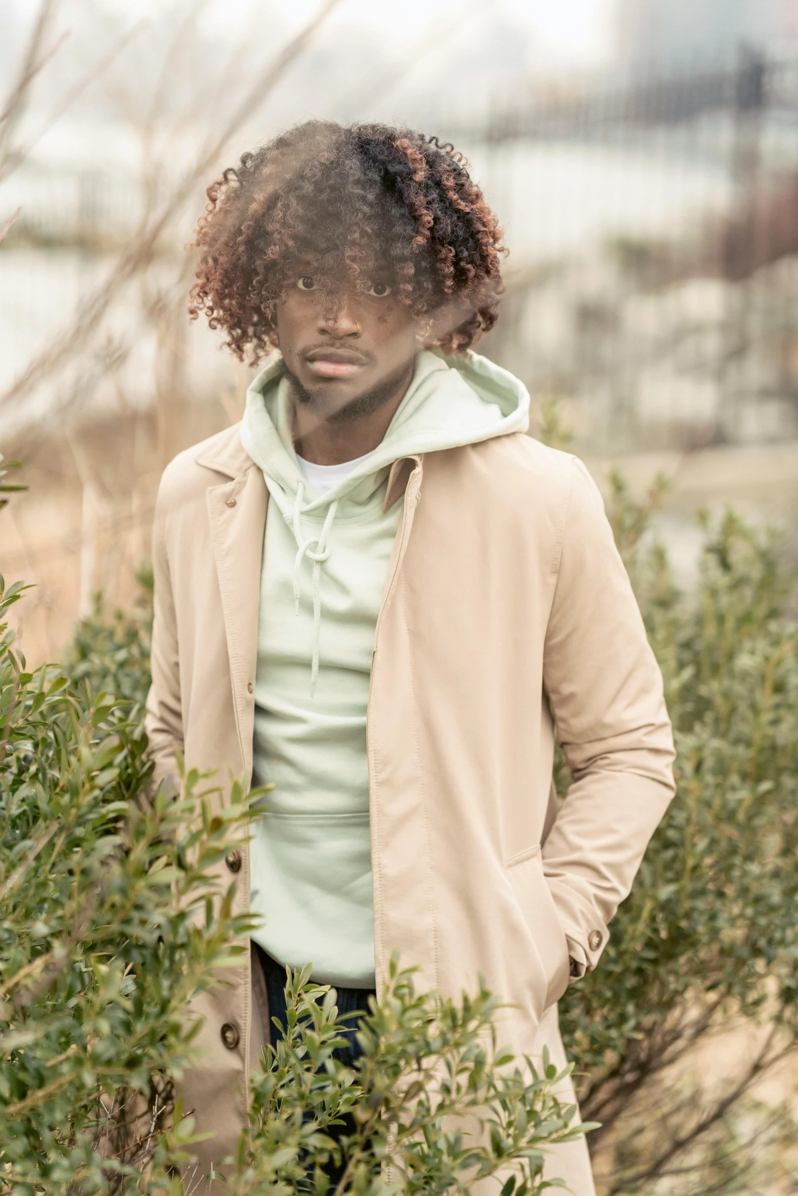 a man wearing a tan jacket is standing behind bushes