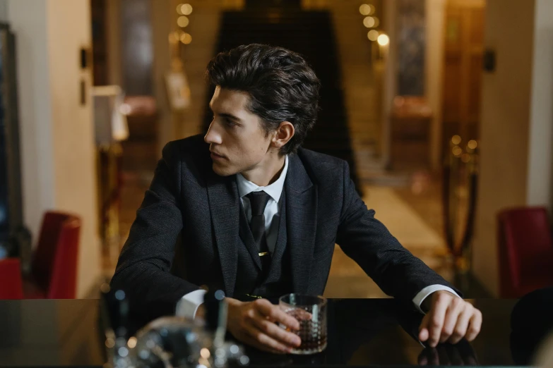 a man in a suit sits at a table with a drink