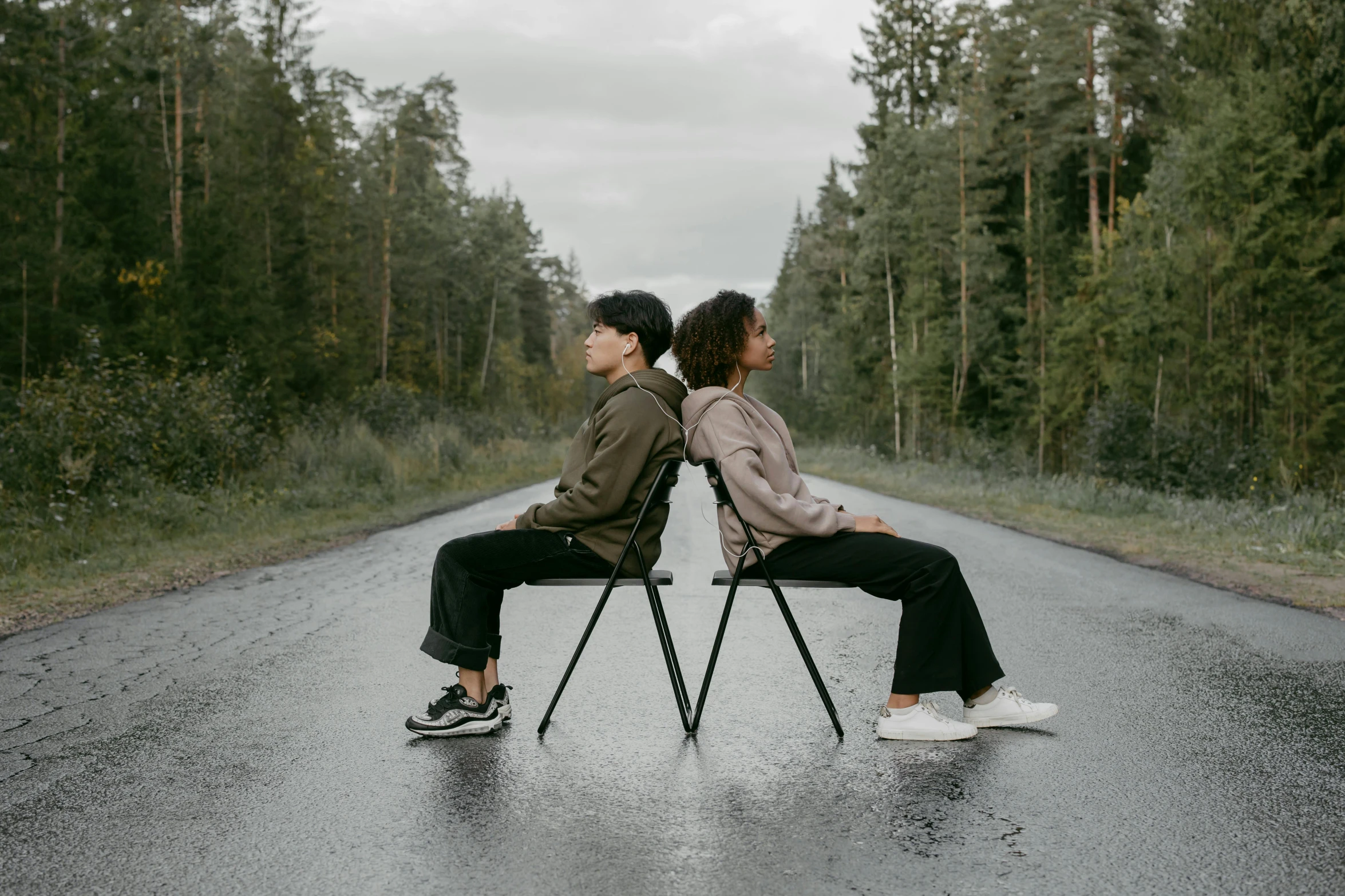 two young people are sitting on a bench on a rainy road