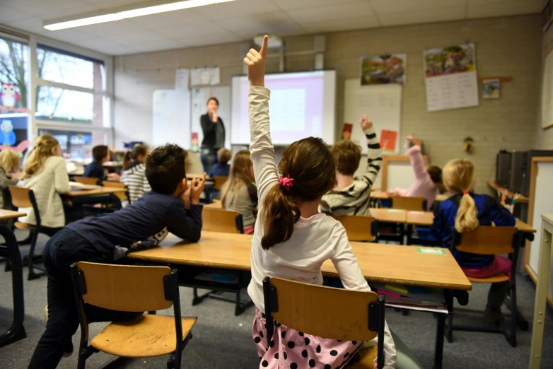 several children sit at desks and wave at the teacher