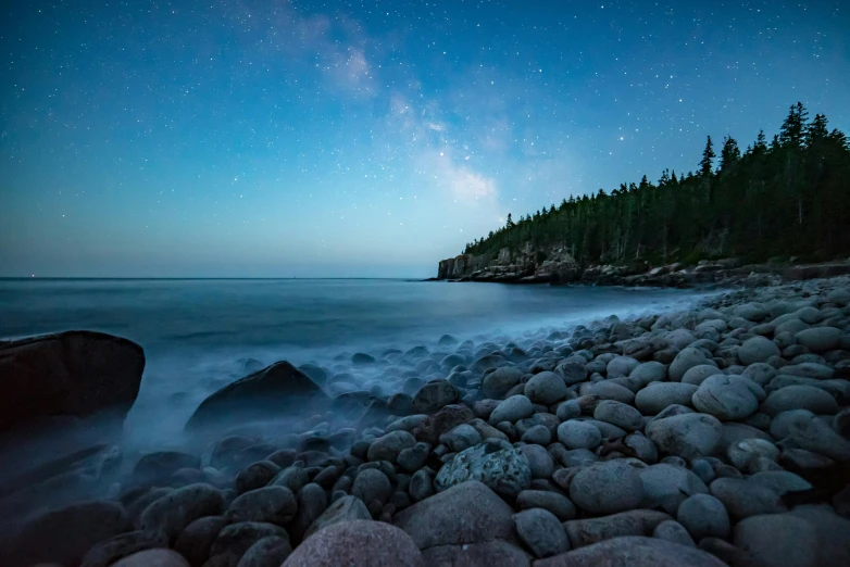 a long exposure of the night sky over a shore with lots of rocks and trees