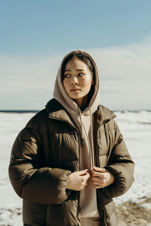 a woman in a hooded coat standing on the beach with her hands under her hair