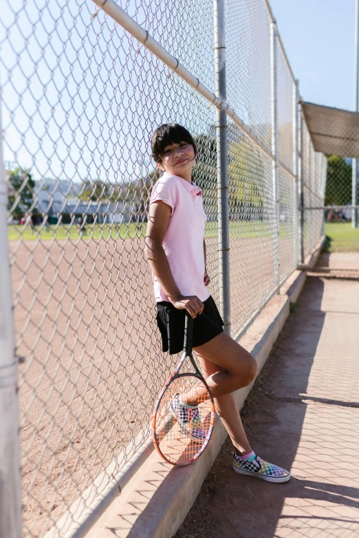 a woman in pink shirt and black shorts leaning on fence with tennis racket