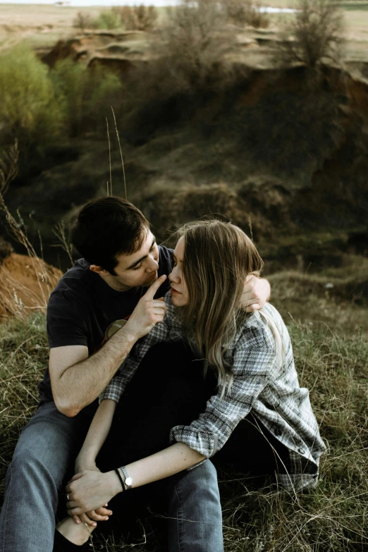 two people sit on the ground, one kissing the other with one eye open