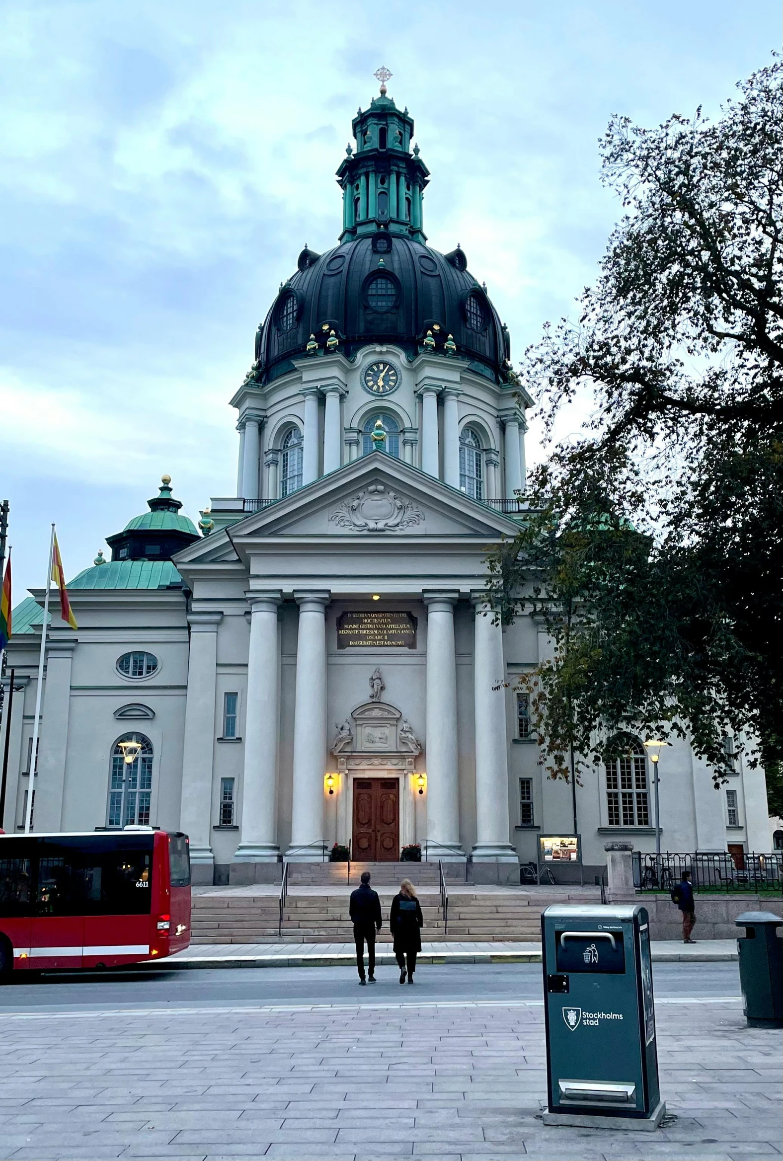 two people walking in front of a white building with two red buses