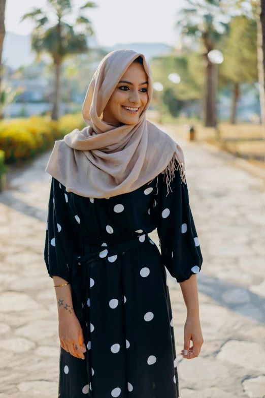 a woman wearing a black dress and a grey and white polka dot hijab is walking down a path