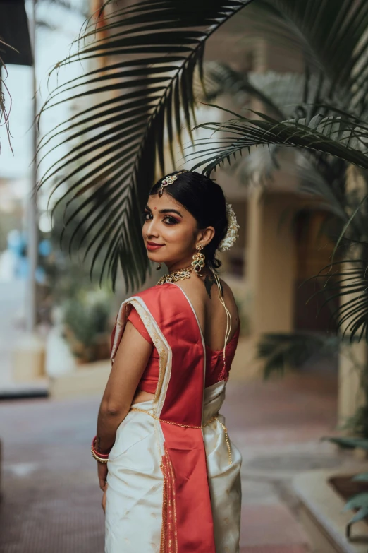 a woman with brown hair wearing a red and white saree standing in front of palm trees