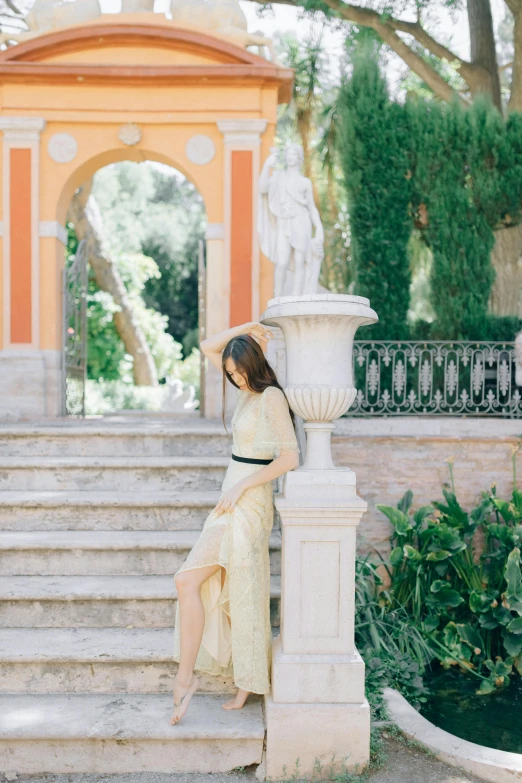 a woman wearing a dress leans on a column in front of a fountain