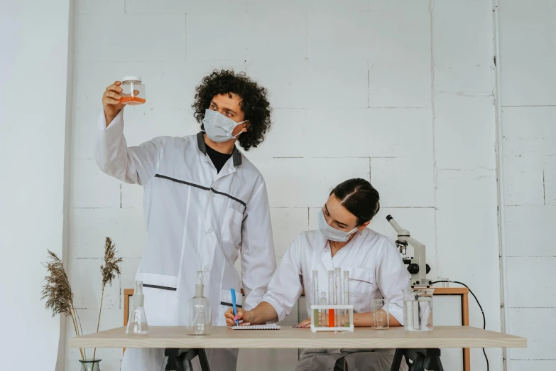 two people in lab coats, one holding a beak and the other wearing a face mask