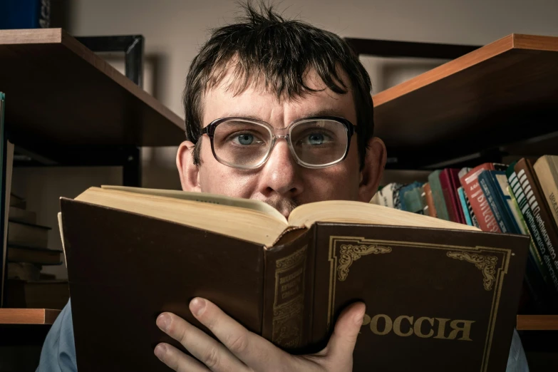a man in glasses and a blue shirt looks through the camera while reading an old book