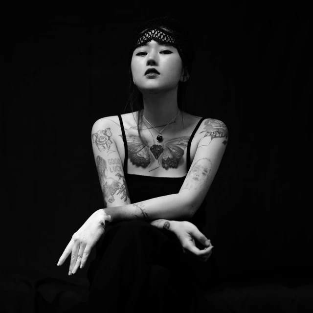 black and white pograph of woman with tattoos on her chest sitting in front of a black background