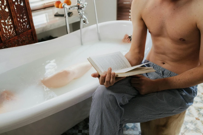 a shirtless man sits in the bathtub holding an open book