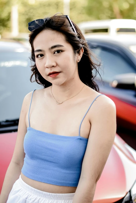 a woman with a bright blue crop top and white skirt stands next to her red car