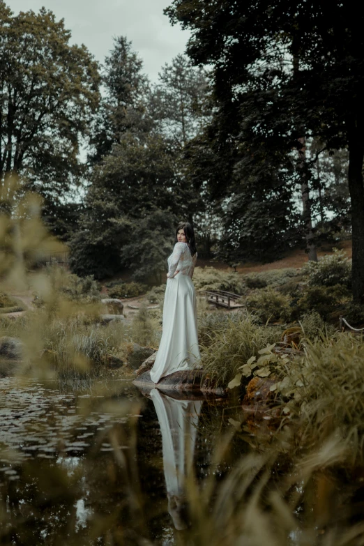 woman in long white dress standing on rock looking into pond