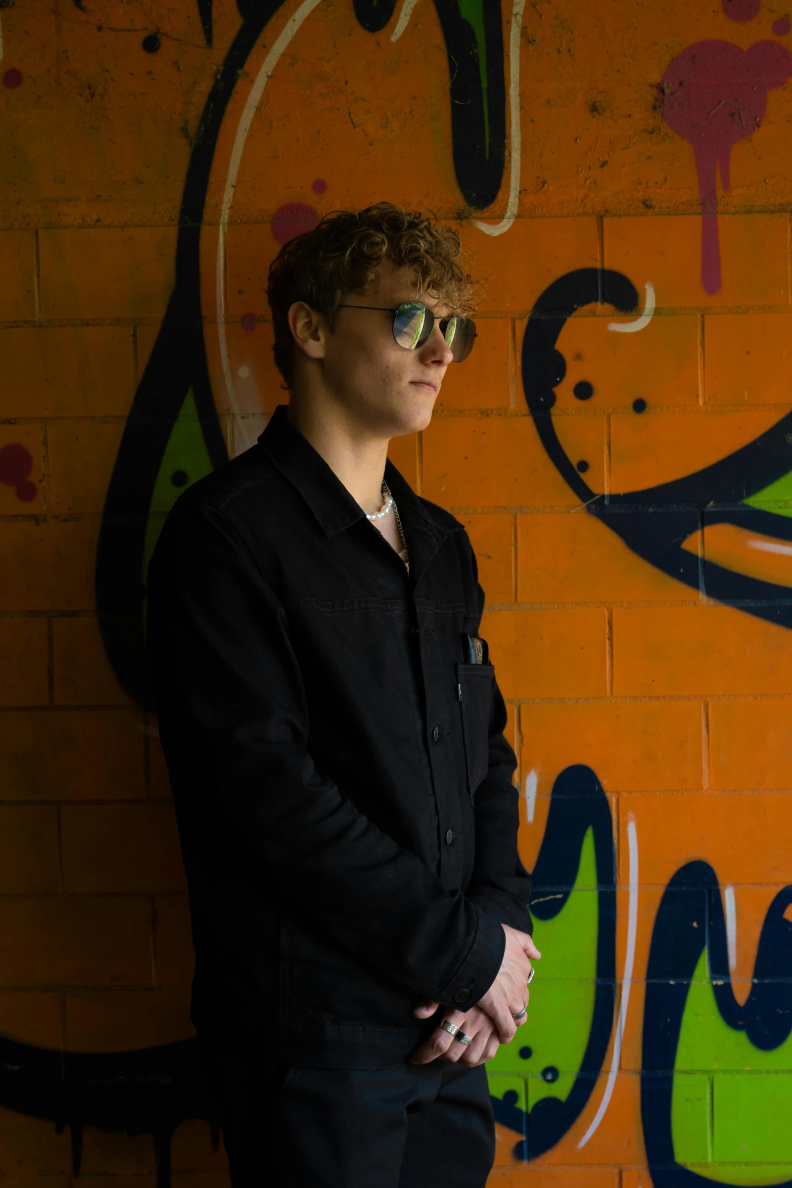 a young man wearing sunglasses stands in front of a wall with graffiti