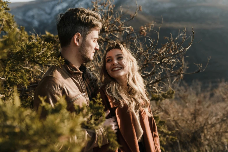 a smiling man standing next to a beautiful blonde woman on a tree covered hillside