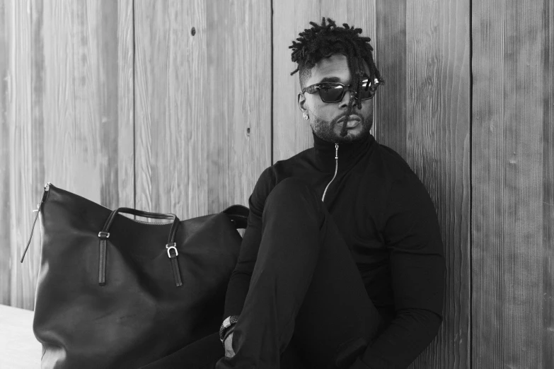 man wearing glasses leaning on wooden wall and holding bag