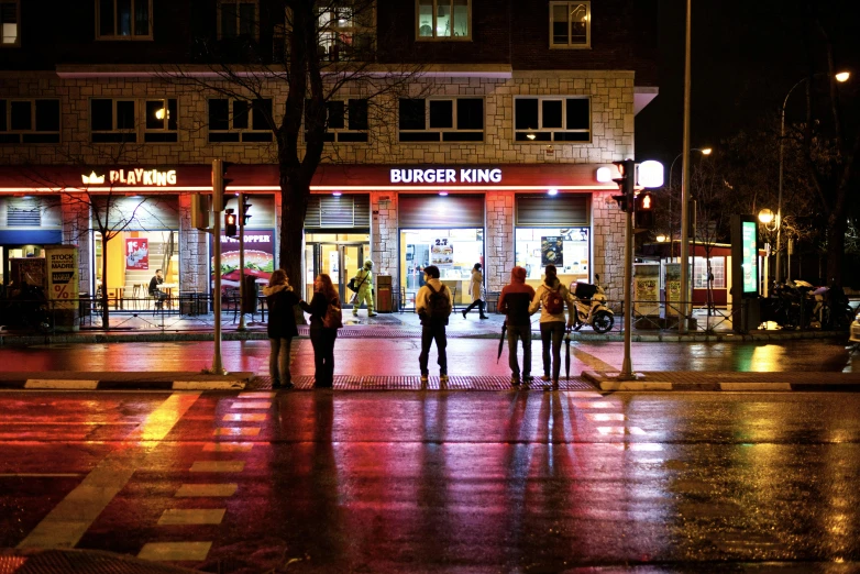 people walking in the rain down a street with a store lit up at night