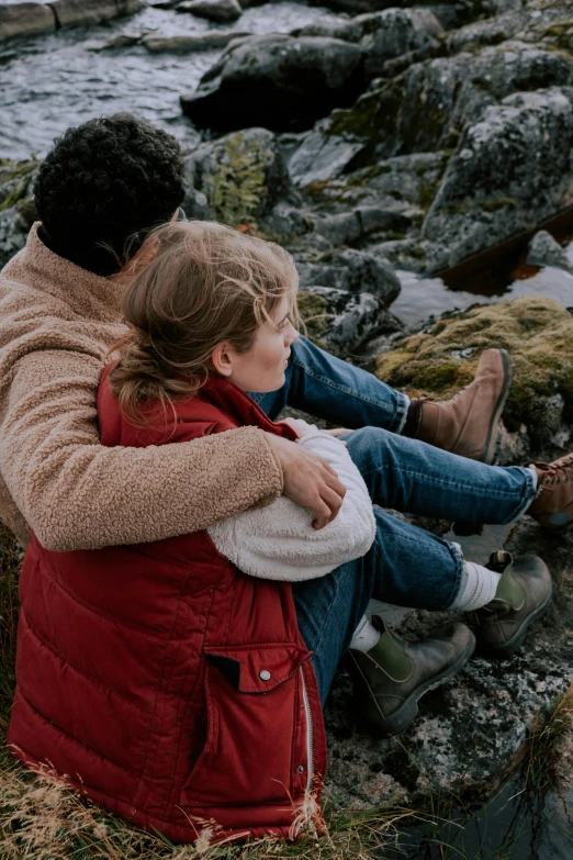 a mother and daughter cuddling on rocks near the water