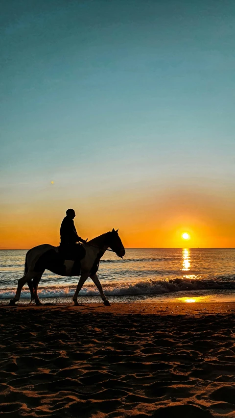 two people ride horses down the beach at sunset