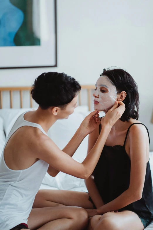 a woman applies on a sheet face mask as another woman sits nearby