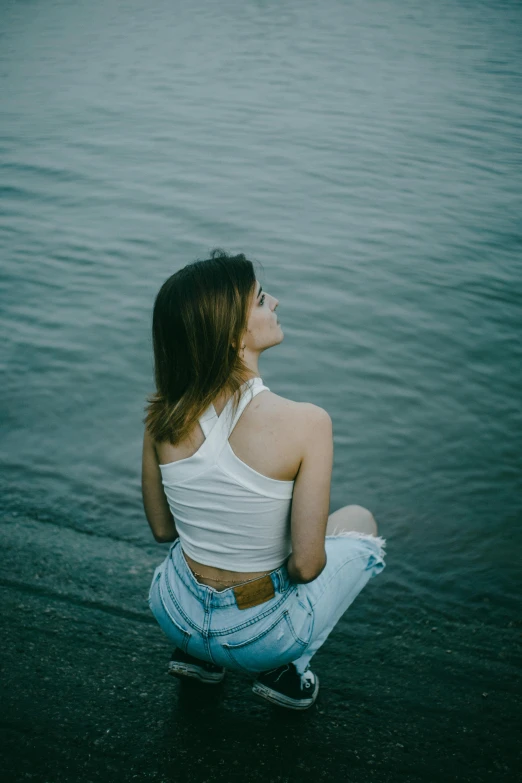 woman in white shirt and blue jeans sitting on skateboard near body of water